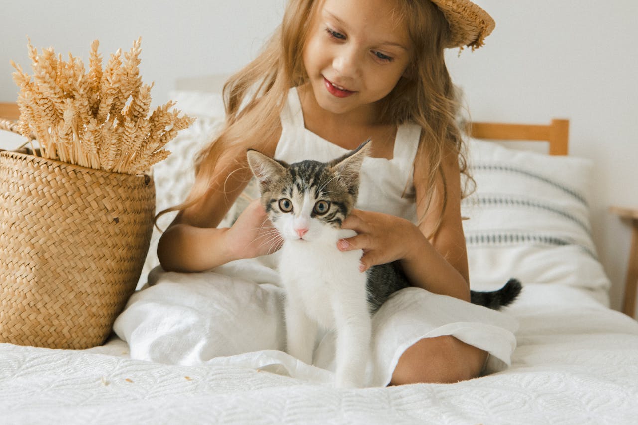 A young girl in a straw hat lovingly pets a kitten on a bed, creating a warm indoor atmosphere.